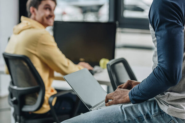 partial view of african american programmer sitting on desk with laptop near smiling colleague