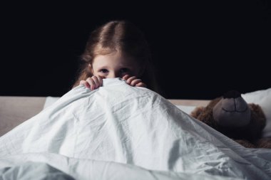 scared child hiding under blanket while sitting on bedding near teddy bear isolated on black clipart