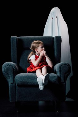 frightened child showing hush sign near white ghost standing behind armchair isolated on black clipart