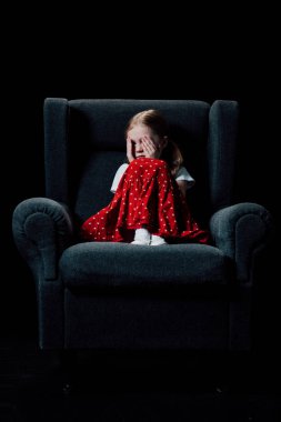 frightened, depressed child sitting in armchair and covering eyes with hands isolated on black clipart