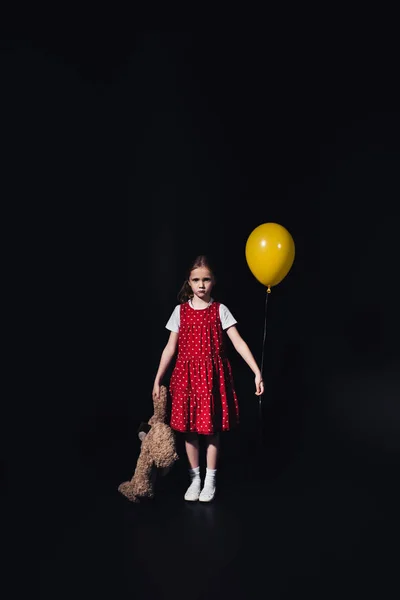 upset, lonely kid with yellow balloon and teddy bear looking at camera isolated on black