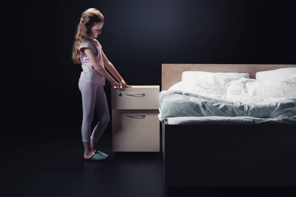 cute child in pajamas opening nightstand on black background