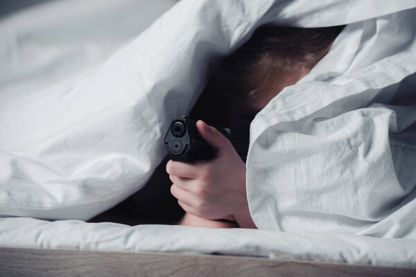 scared child holding gun while hiding under blanket in bedroom