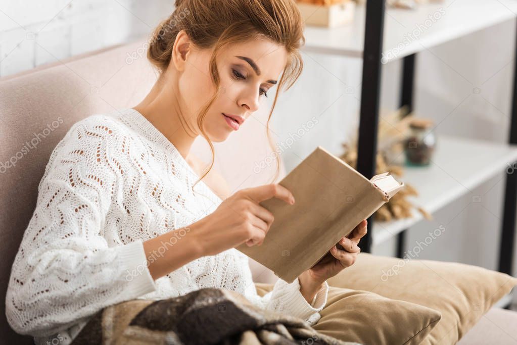 attractive woman in white sweater sitting on sofa and reading book 
