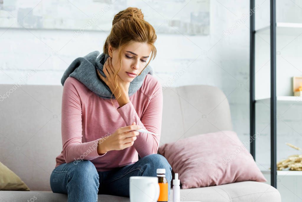 attractive and ill woman with grey scarf looking at thermometer 