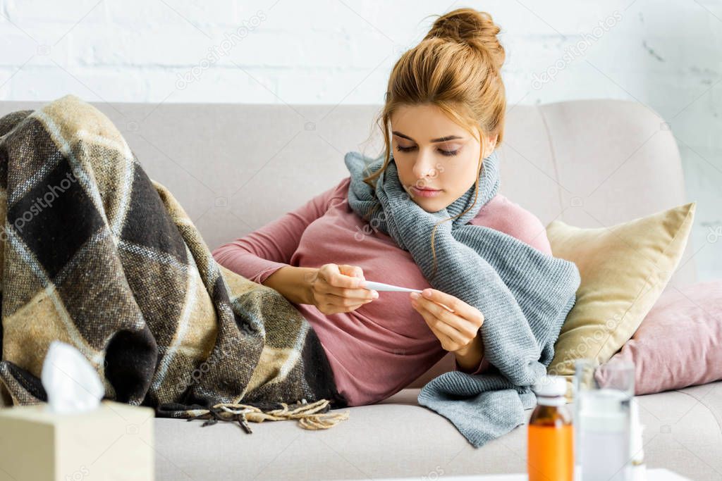 attractive and ill woman with grey scarf looking at thermometer 