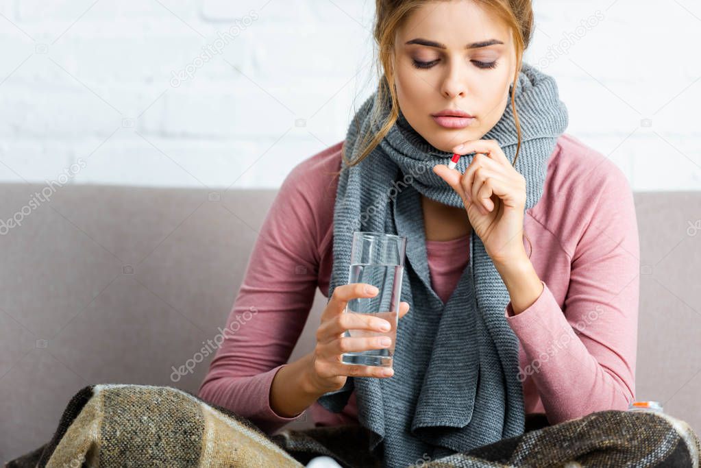 attractive and ill woman with grey scarf taking pill and holding glass of water 