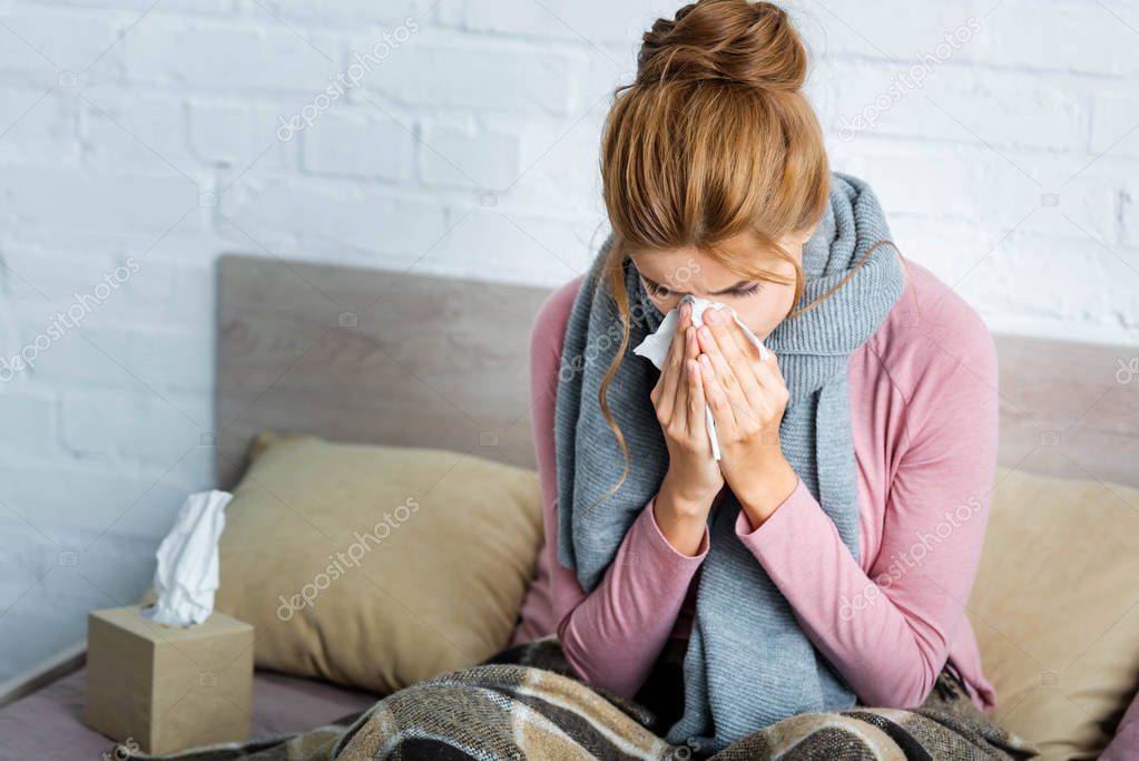 attractive and ill woman with grey scarf sneezing and using napkin