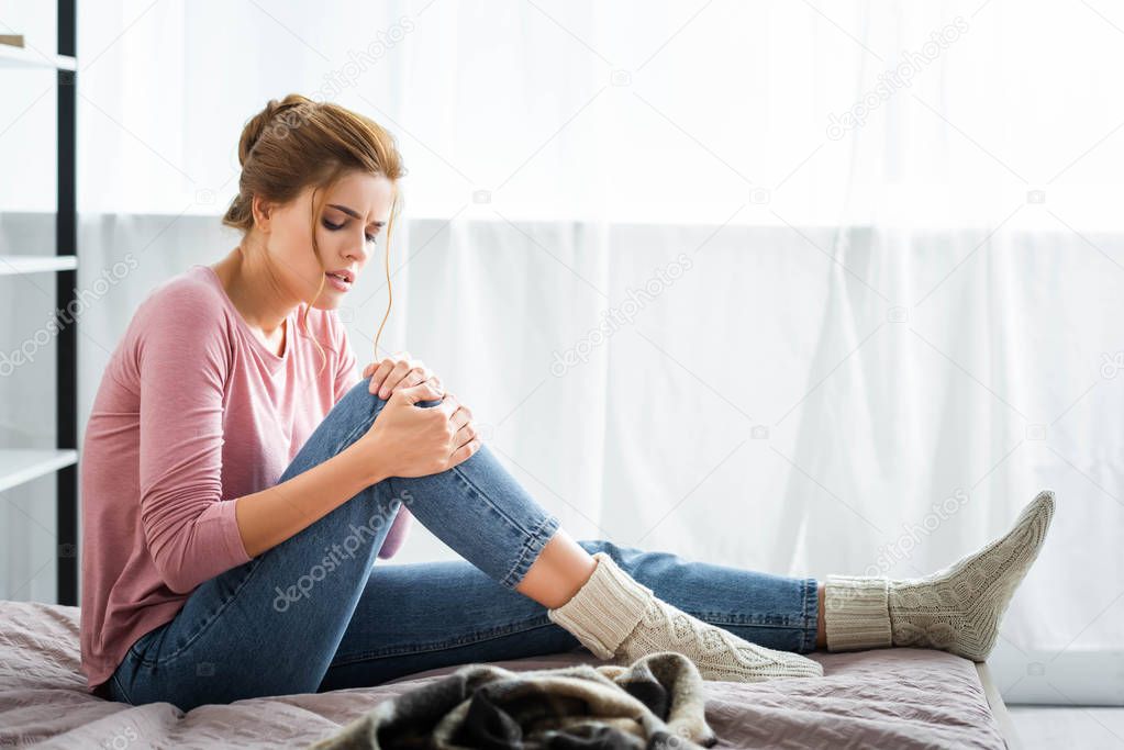 attractive woman with pain in knee sitting on bed in apartment 