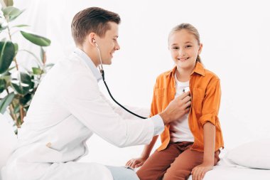 stetoskoplu çocuğu inceleyen beyaz önlüklü gülümseyen pediatrist