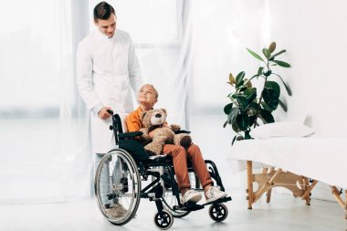 beyaz önlük ve tekerlekli sandalyede oyuncak ayı ile çocuk pediatrist tam uzunlukta görünümü