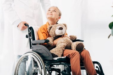 beyaz önlük ve tekerlekli sandalyede oyuncak ayı ile çocuk pediatrist kırpılmış görünümü