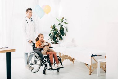 beyaz önlük ve tekerlekli sandalyede oyuncak ayı ile çocuk pediatrist tam uzunlukta görünümü