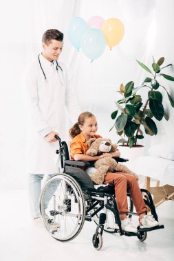 beyaz önlük ve tekerlekli sandalyede oyuncak ayı ile çocuk pediatrist tam uzunlukta görünümü