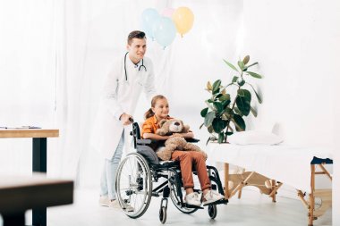 beyaz önlük ve tekerlekli sandalyede oyuncak ayı ile çocuk pediatrist tam uzunlukta görünümü