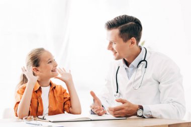 gülümseyen çocuk ve pediatrist klinikte masada otururken birbirlerine bakıyor
