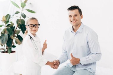 doktor kostümü ve hasta el sıkışma ve başparmak gösteren gülümseyen çocuk
