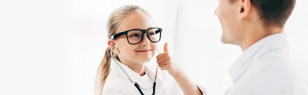 panoramic shot of smiling kid in doctor costume looking at patient and showing thumb up