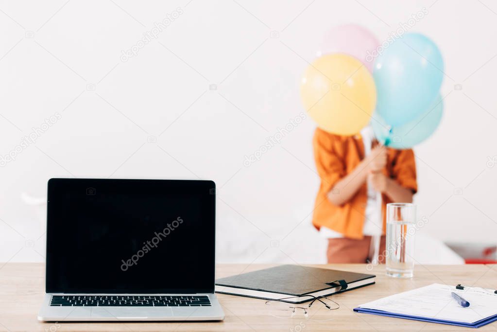 selective focus of kid holding colorful balloons and laptop with blank screen on foreground