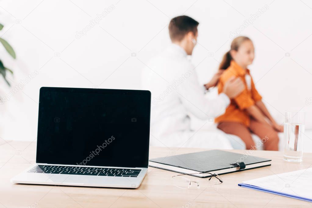 selective focus of pediatrist examining child and laptop with blank screen on foreground