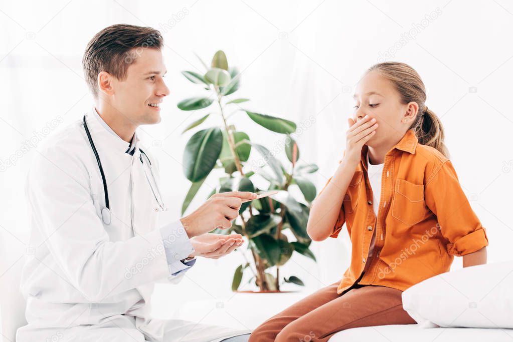kid covering mouth with hand and smiling pediatrist holding medical spatula in clinic