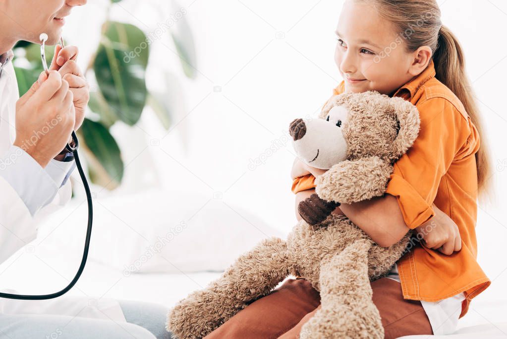 partial view of pediatrist in white coat and kid with teddy bear in clinic