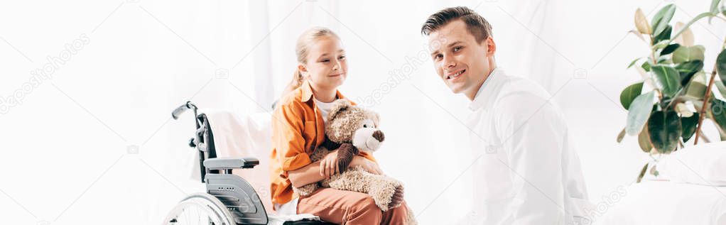 panoramic shot of pediatrist and kid with teddy bear on wheelchair