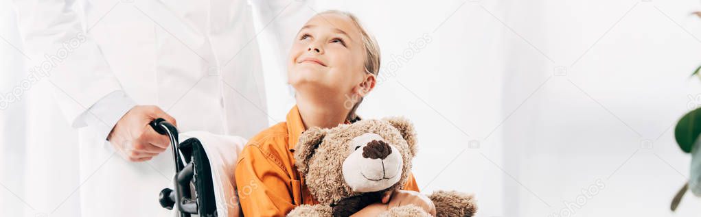 panoramic shot of pediatrist in white coat and kid with teddy bear on wheelchair