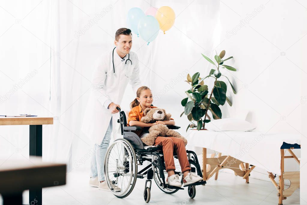 full length view of pediatrist in white coat and kid with teddy bear on wheelchair