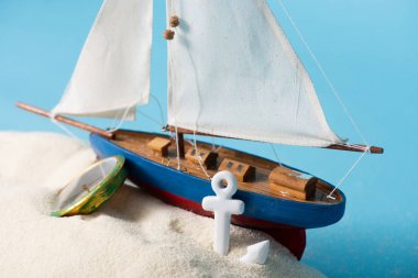 miniature ship near compass and anchor in white sand isolated on blue clipart
