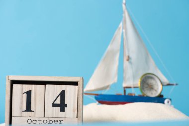 wooden calendar with 14 October date near decorative ship and compass  in white sand isolated on blue clipart