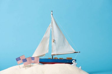 decorative ship near American national flags in white sand isolated on blue clipart