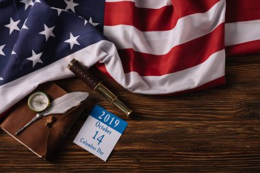 top view of calendar with Columbus Day inscription near leather notebook, nib, telescope and compass on wooden surface with American national flag clipart