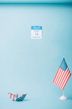 paper calendar with Columbus Day inscription near paper boat and American national flag on blue background clipart