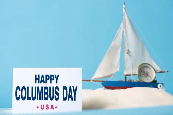 青で隔離された白い砂のミニチュア船の近くに幸せなコロンブスの日の碑文を持つカード — ストック写真