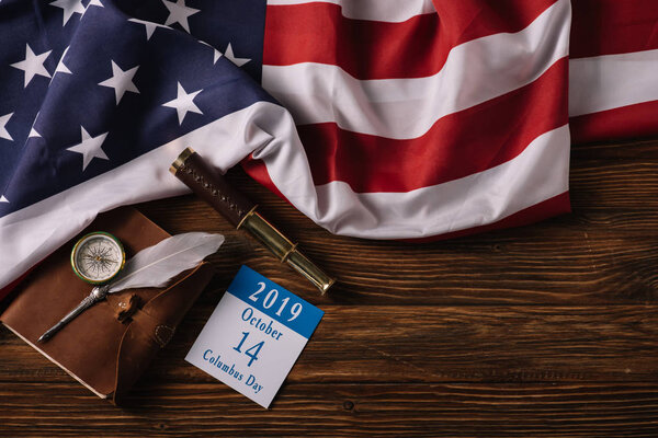 top view of calendar with Columbus Day inscription near leather notebook, nib, telescope and compass on wooden surface with American national flag