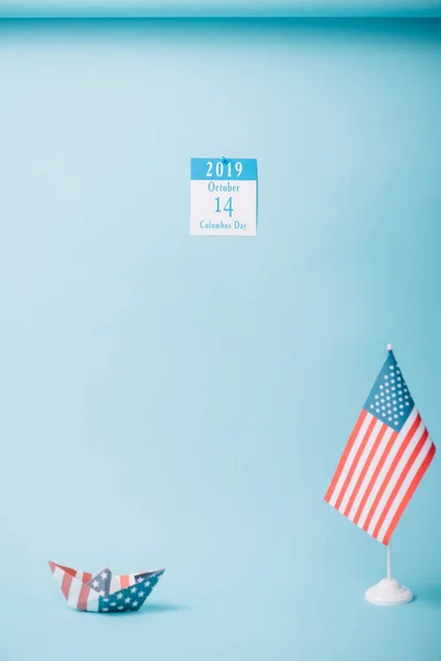 paper calendar with Columbus Day inscription near paper boat and American national flag on blue background