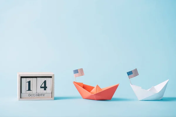 wooden calendar with October 14 date near paper boats with American flag pattern on blue background 