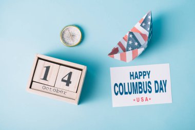 top view of calendar with October 14 date near paper boat with American flag pattern, compass and card with happy Columbus Day inscription on blue background  clipart