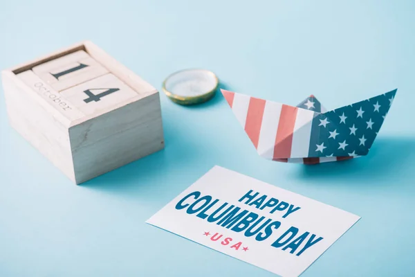 wooden calendar with October 14 date near paper boat with American flag pattern, compass and card with happy Columbus Day inscription on blue background 