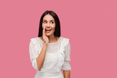 cheerful woman touching face while gossiping isolated on pink  clipart