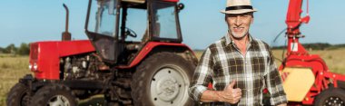 panoramic shot of happy self-employed farmer in straw hat showing thumb up near tractor  clipart