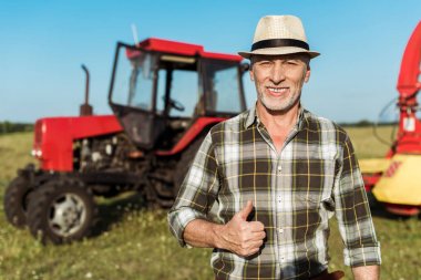 happy farmer in straw hat showing thumb up near tractor  clipart