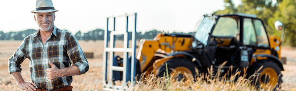 panoramic shot of happy bearded farmer showing thumb up near tractor 