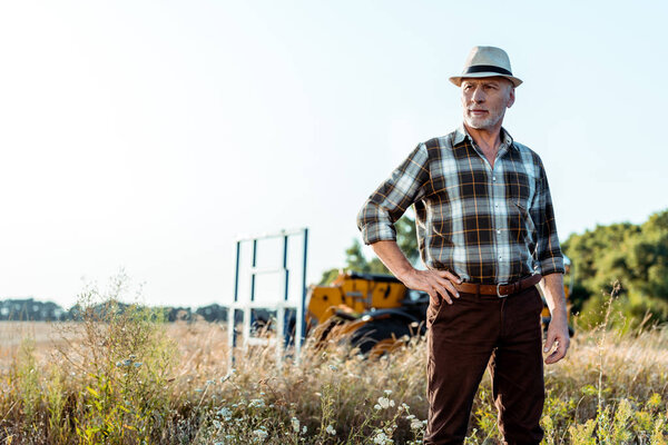 senior man in straw hat standing with hand on hip near tractor 
