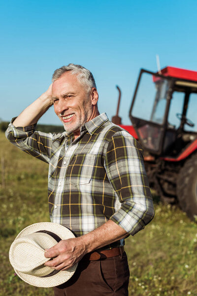 happy self-employed farmer holding straw hat near tractor 