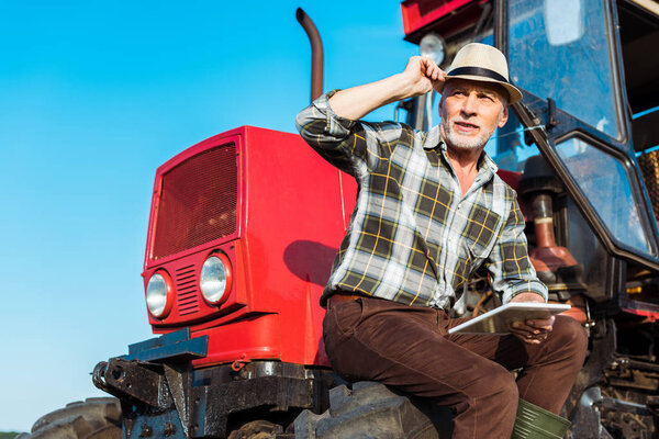 senior farmer in straw hat holding digital tablet near red tractor 