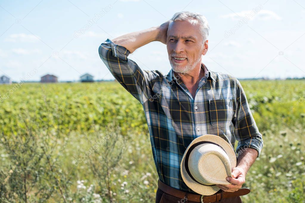 smiling senior farmer holding straw hat in field 