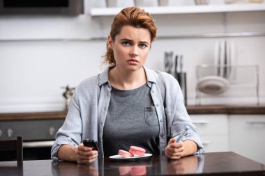 tatlı alerjisi olan üzgün kadın pembe tatlı yanında oturan ve glikoz monitör tutan