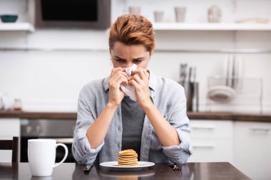 sick woman sneezing in napkin near tasty pancakes at home  clipart